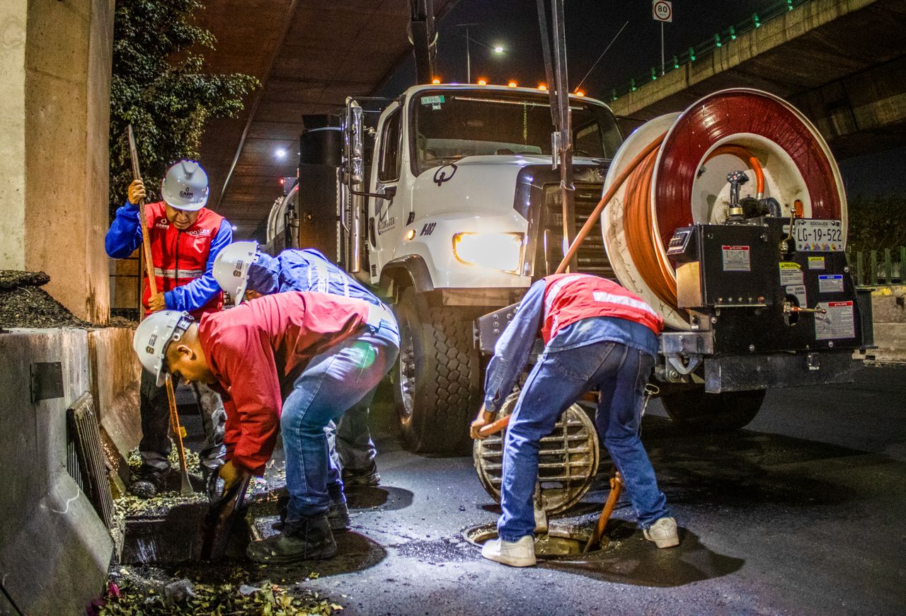 our-story-01 Street workers cleaning drainage at night with machinery under a bridge.